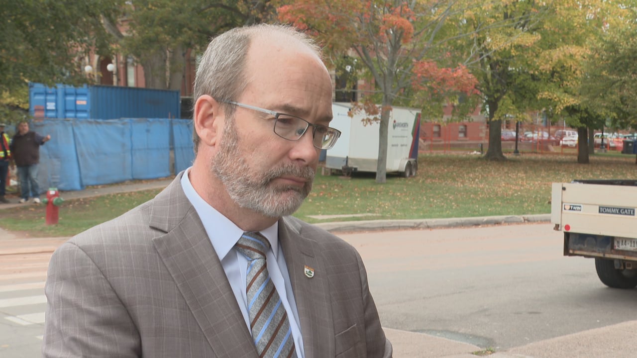 A man in a suit stands outside a building near a street. He is not looking at the camera. 
