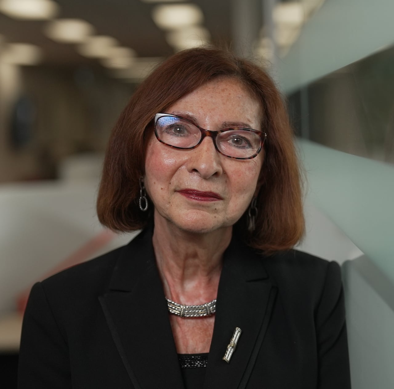 A woman with shoulder length auburn hair wearing glasses leans against a glass wall as she looks into the camera.