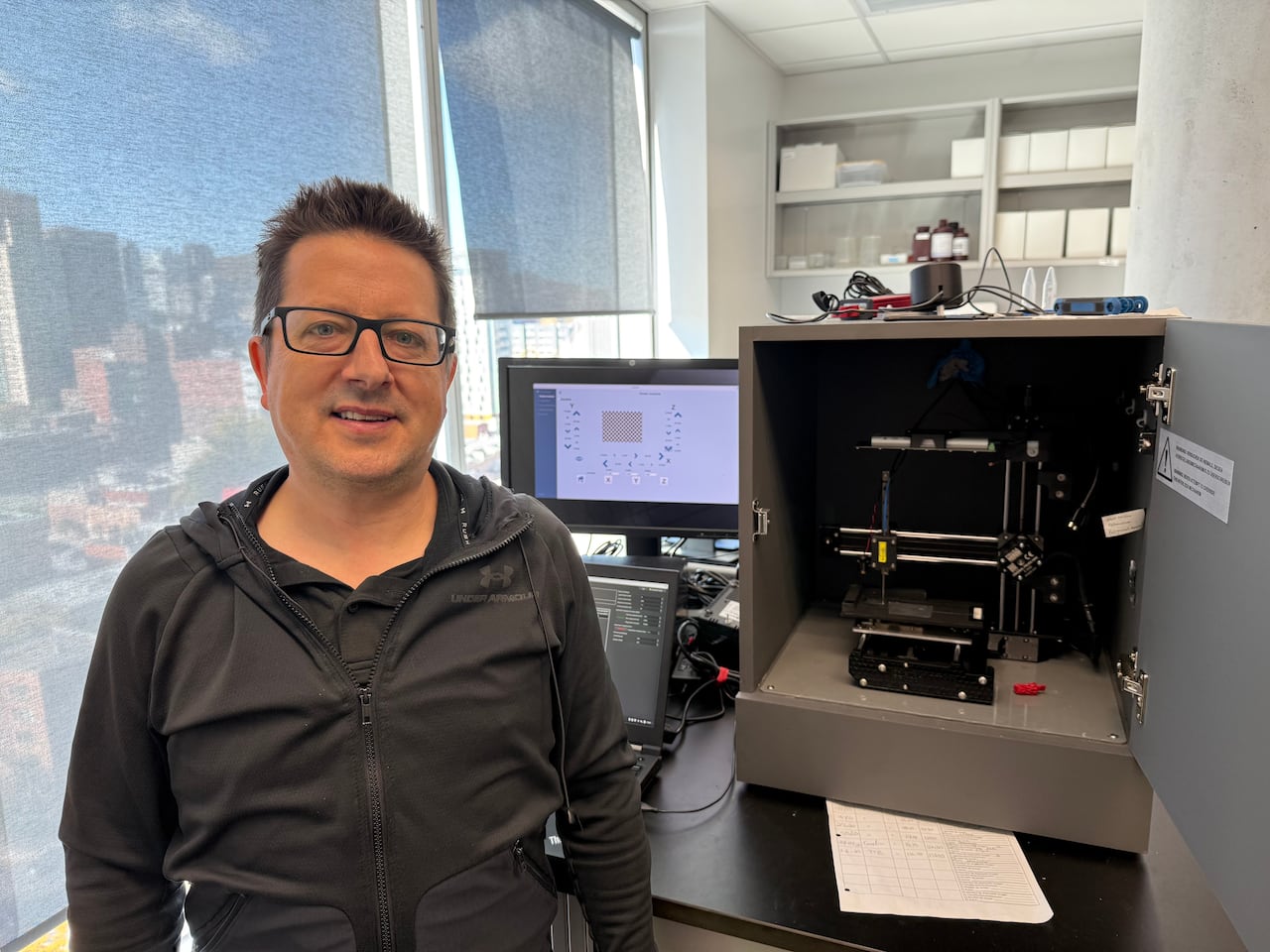 A man with black glasses stands next to a machine with a needle.