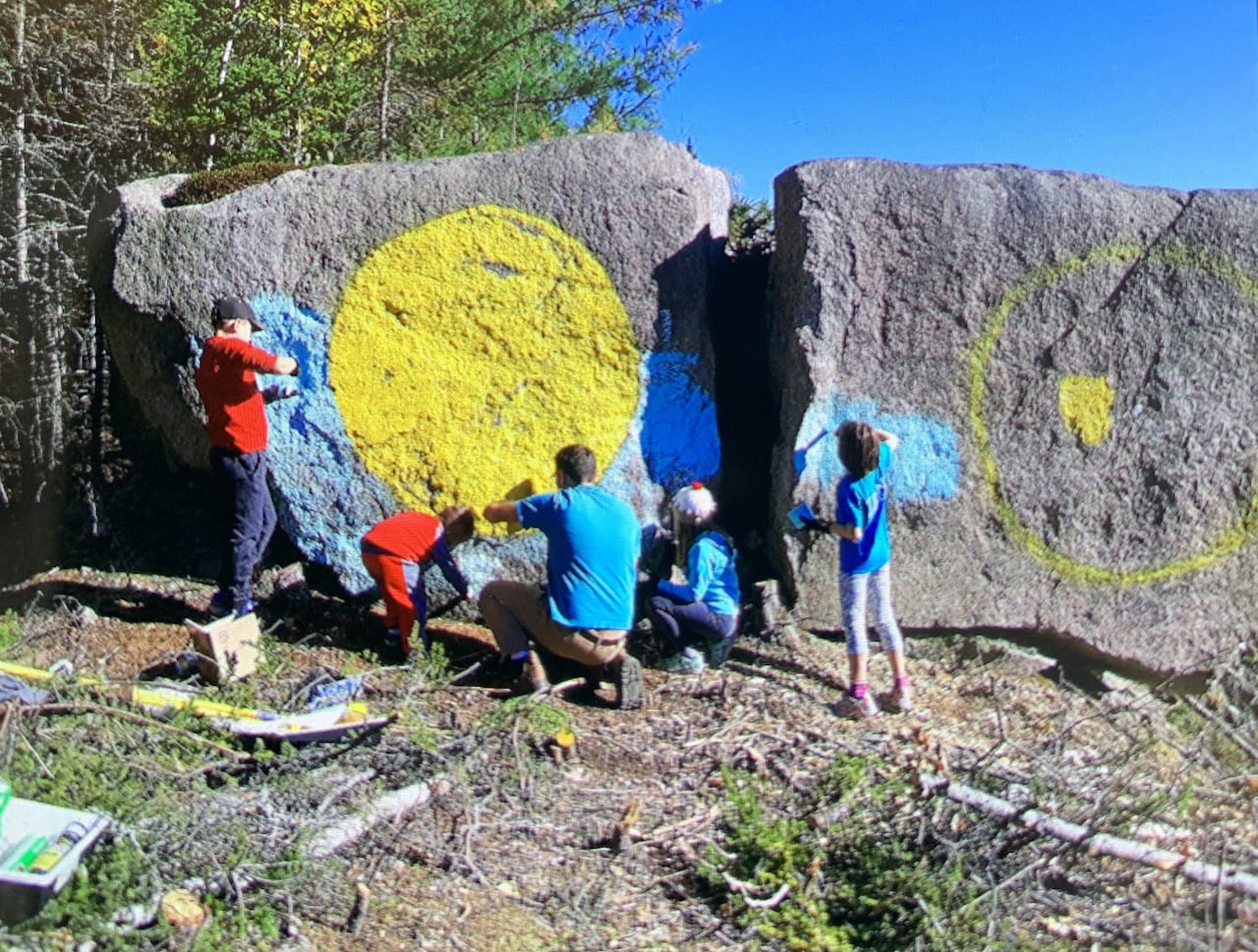 A group of people paint a couple large boulders on a nice day.