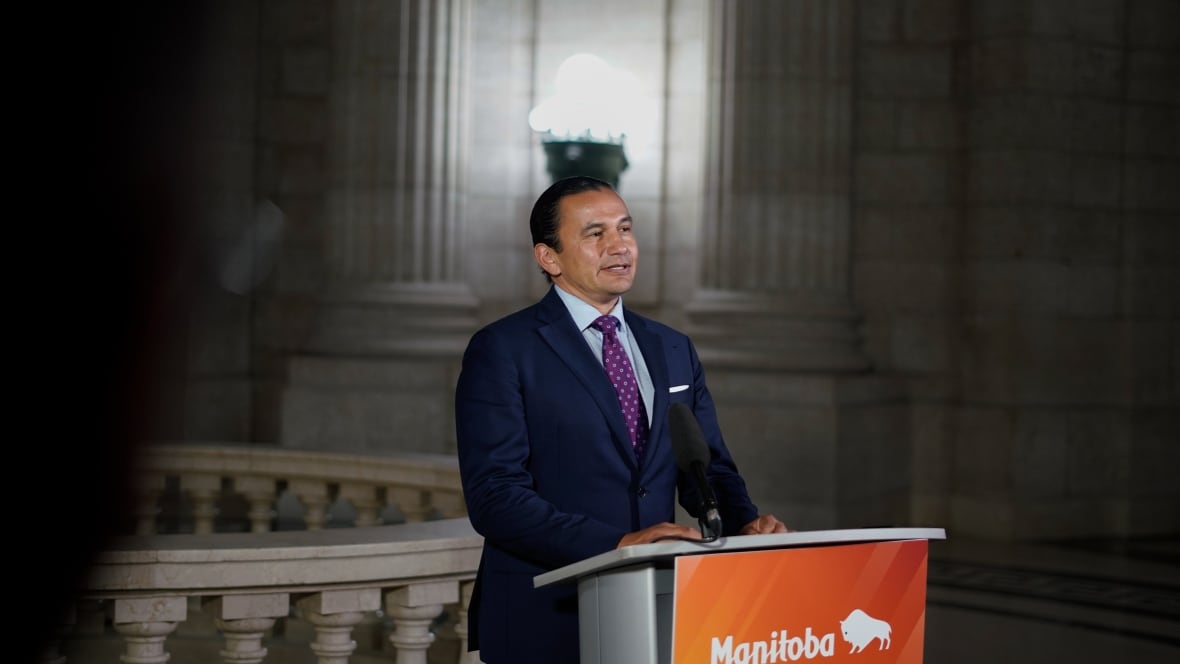 A man in a dark blue suit, purple tie and dark hair tied back in a pony tail stands in front of a lectern and speaks into a microphone to media.