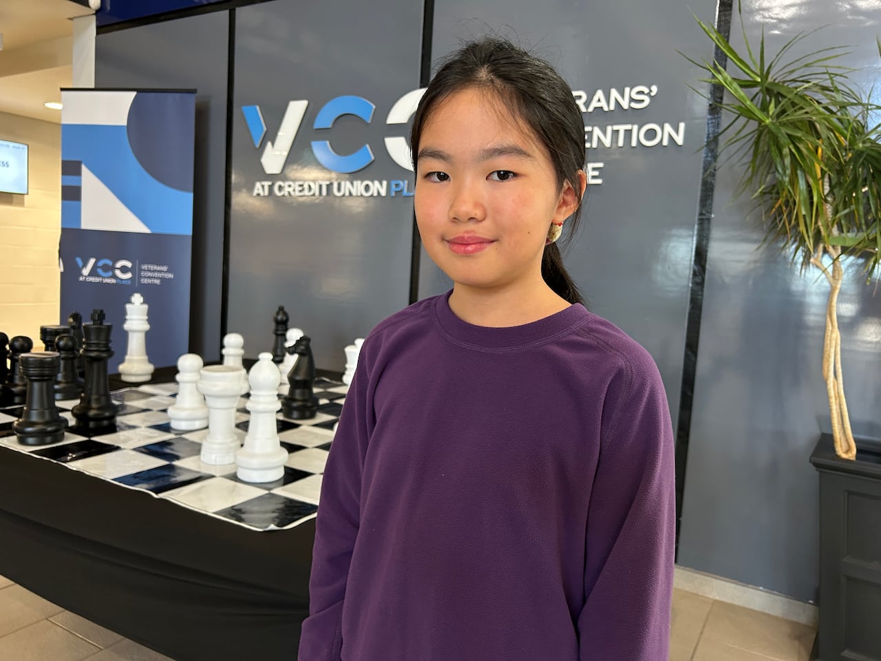 A young girl with black hair is wearing a purple sweater. Behind her is an oversized chess board.