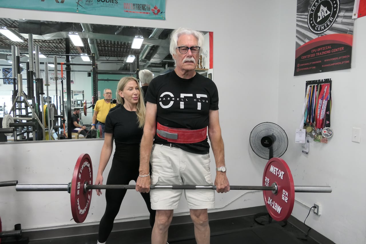 A man with white hair and a white moustache lifts a bar with two red 25 kg weights on either side at a gym. A woman in gym clothes stands behind, spotting him.