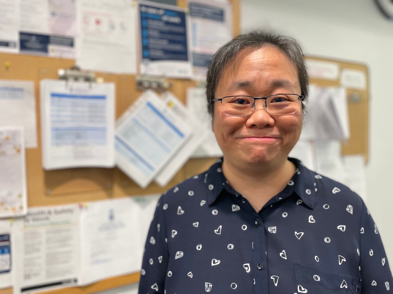 A woman smiles into the camera. She is wearing a dark blouse with hearts on it. She is standing in front of a corkboard