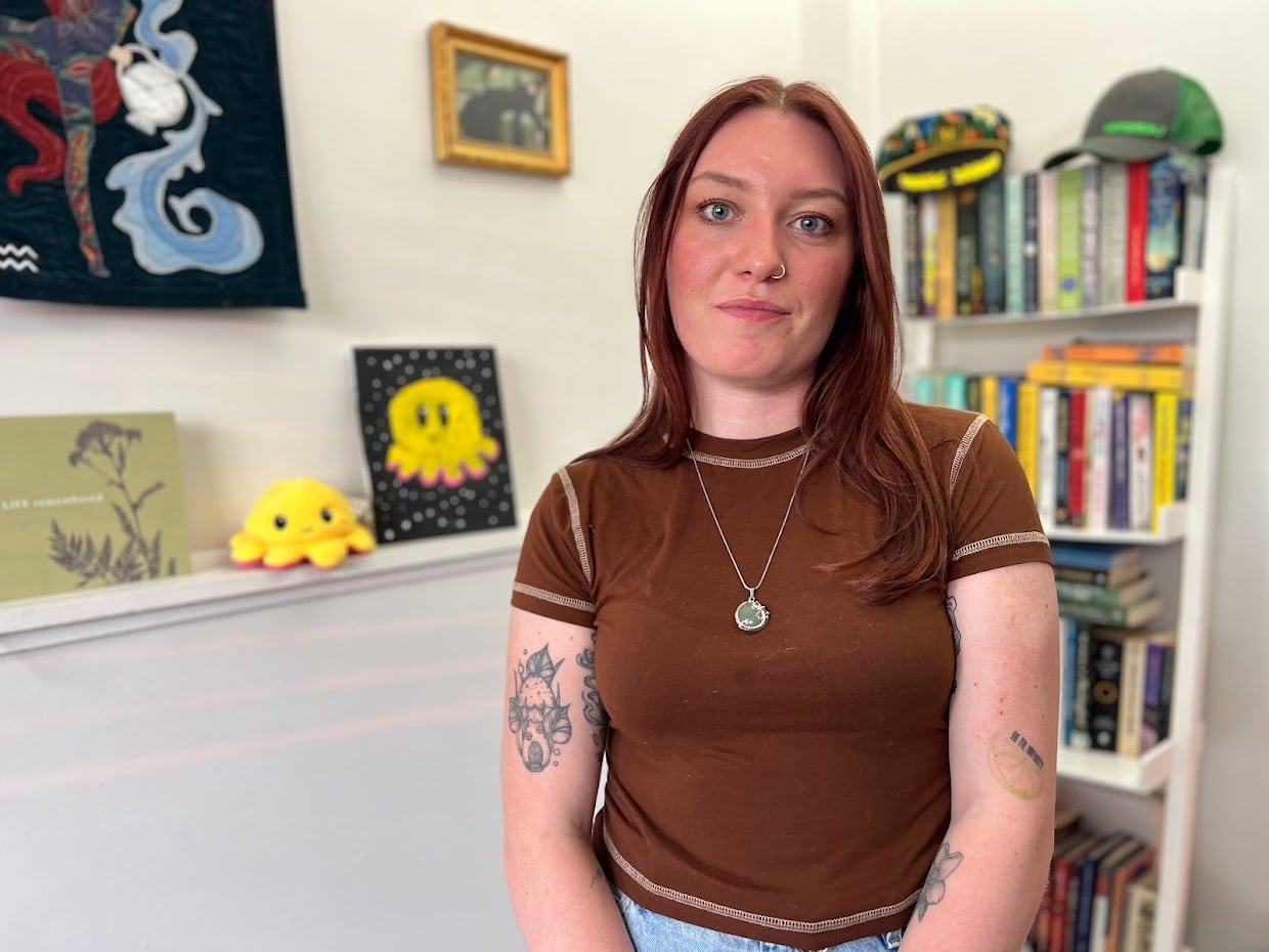 A woman with long reddish hair and 4 tattoos in a brown t-shirt and silver  necklace stands in front a wall with paintings and bookshelf behind her. 