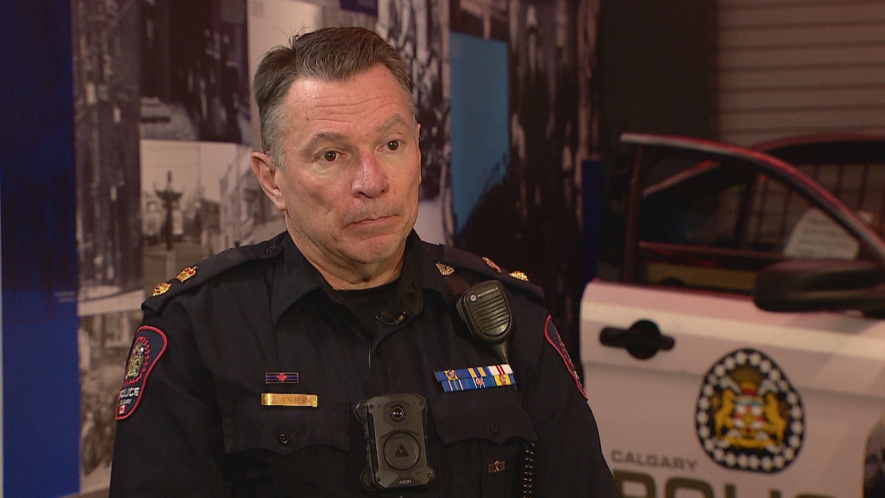 A man with short dark hair that's greying at the sides is wearing  a police uniform sitting in front of a police cruiser with open doors.  