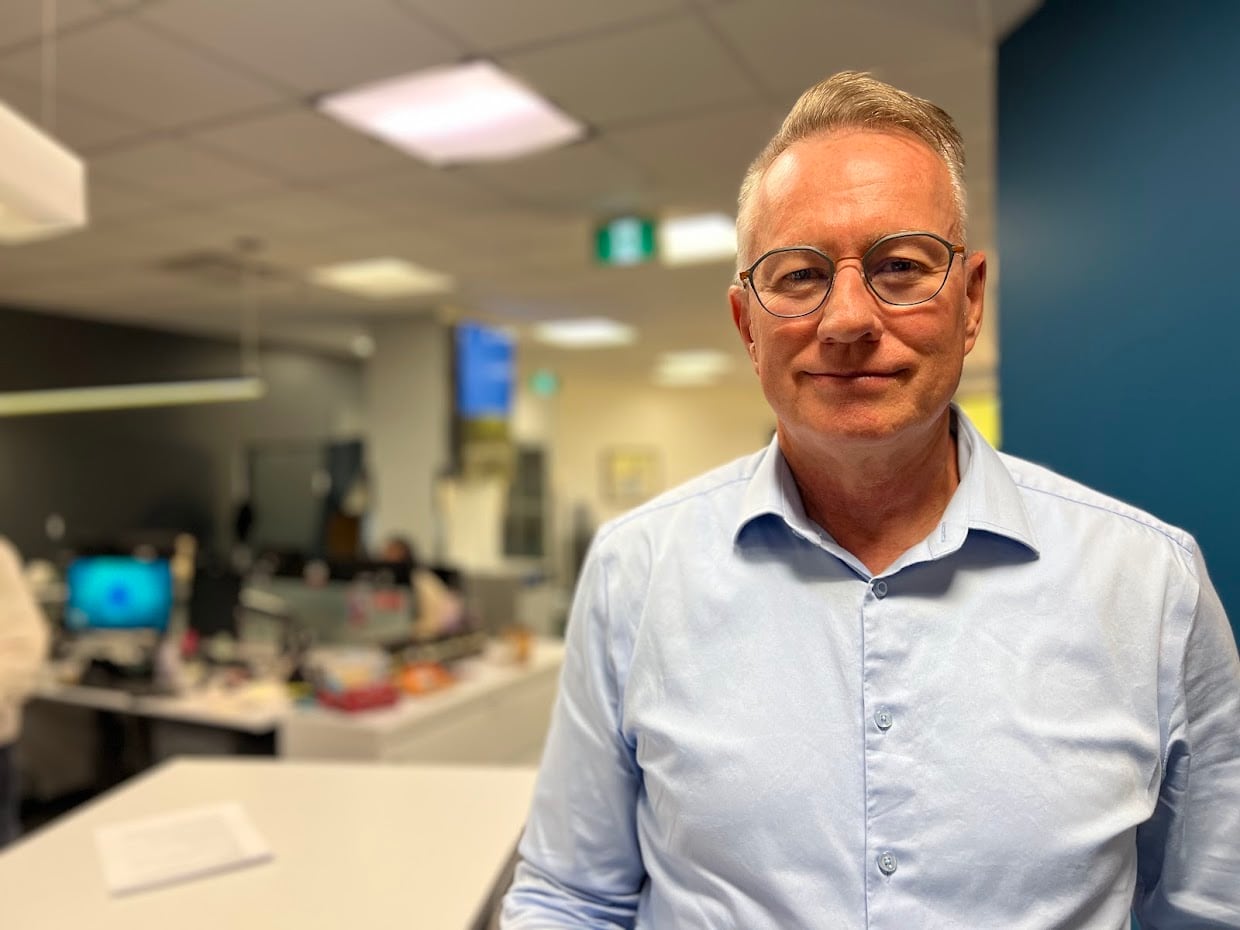 A middle aged man in a light blue dress shirt with grey hair and glasses stands in an open concept office space. 