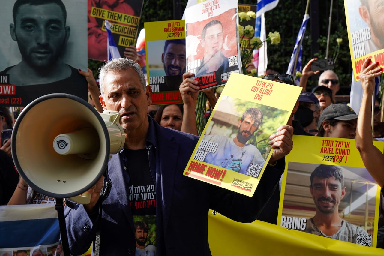 A white-haired man is shown holding up a sign bearing the picture of a younger, bearded man, at what appears to be an outdoor demonstration.