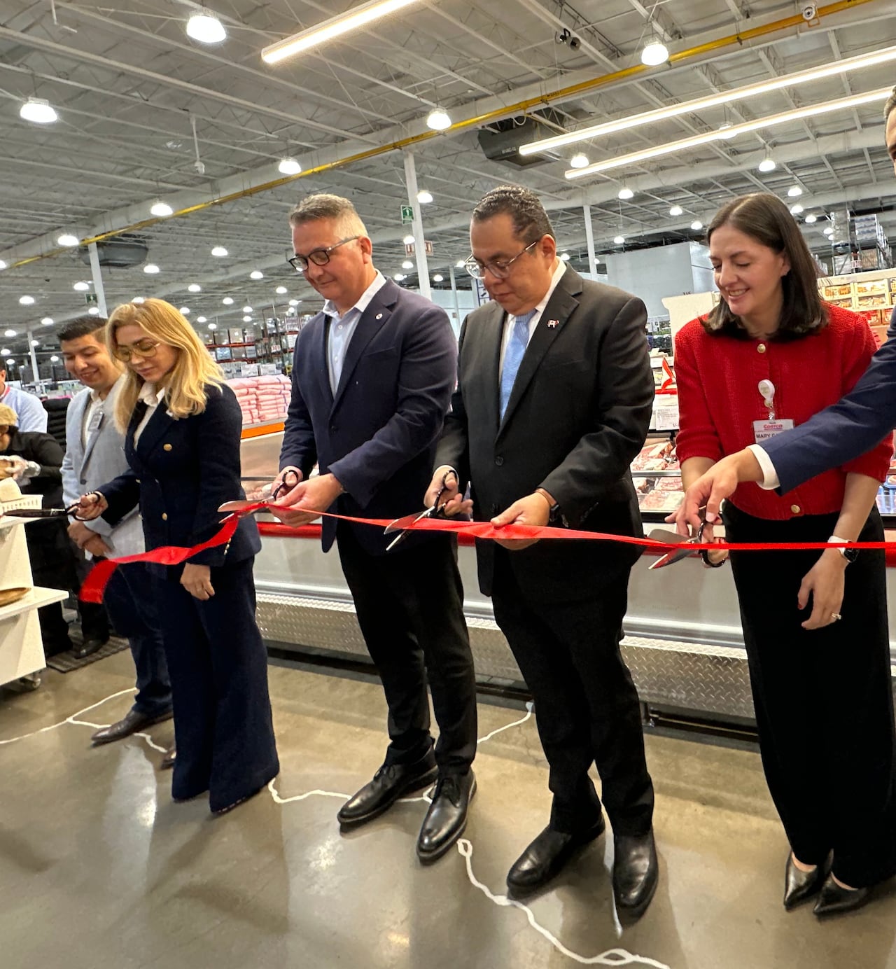 A line of people stand with a red ribbon and scissors in hand by a cooler full of beef products.