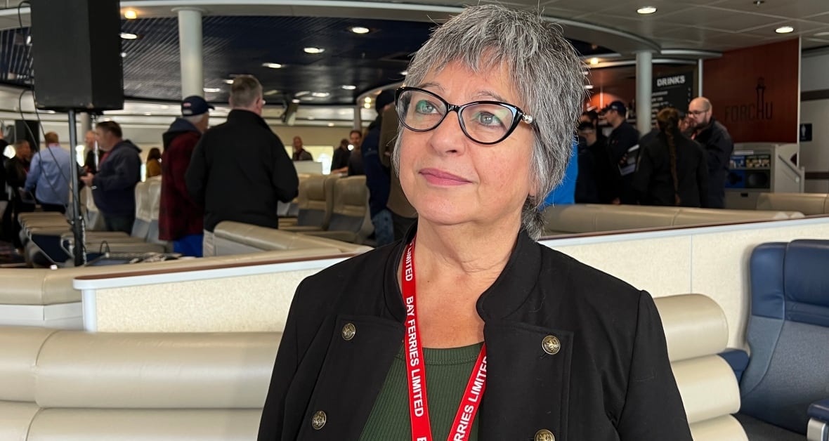 A woman with short grey hair and glasses wearing a blazed and a Bay Ferries lanyard stands on a ferry with people mingling behind her.