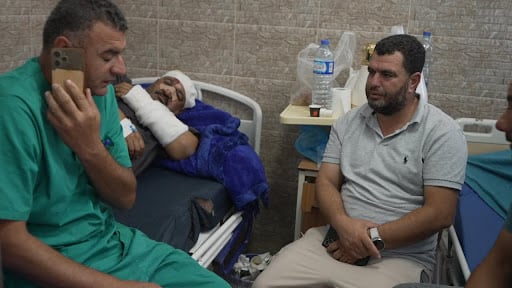 Dr. Tareq Abu Aram sits on a hospital bet with a man in an arm cast and head bandage behind him. 