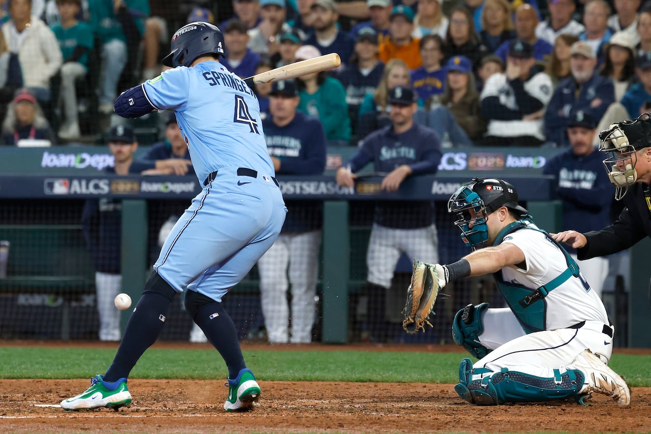 A man in a baseball uniform stands with his back to the camera at home plate as a baseball comes at his knee. A catcher holds out his glove behind the batter. 