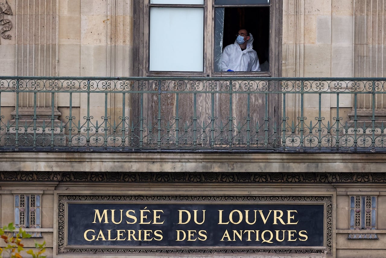 A man in a white suit and mask looks through a broken window.
