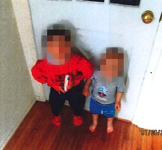 Two boys stand in a doorway. The older boy is wearing a Spiderman costume. 