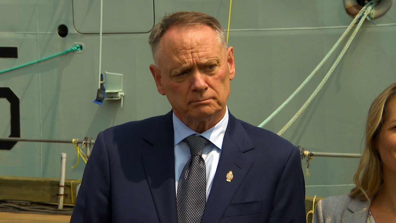 A man stands in front of a new naval ship while taking questions from reporters. 