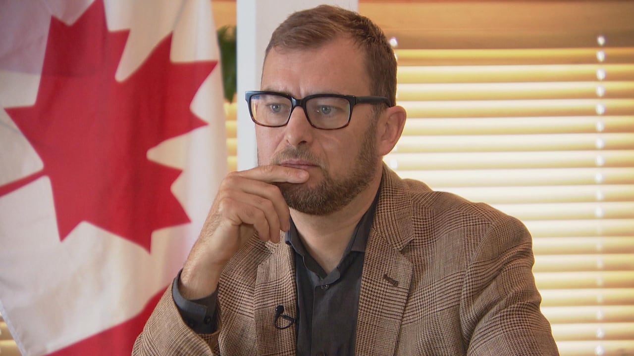 A bearded man in a brown suit sits with his chin in his hand.