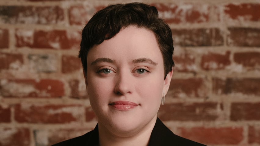 A portrait of a smiling young woman with short, dark hair, wearing a cream-coloured blouse and black blazer, standing in front of a brick wall.
