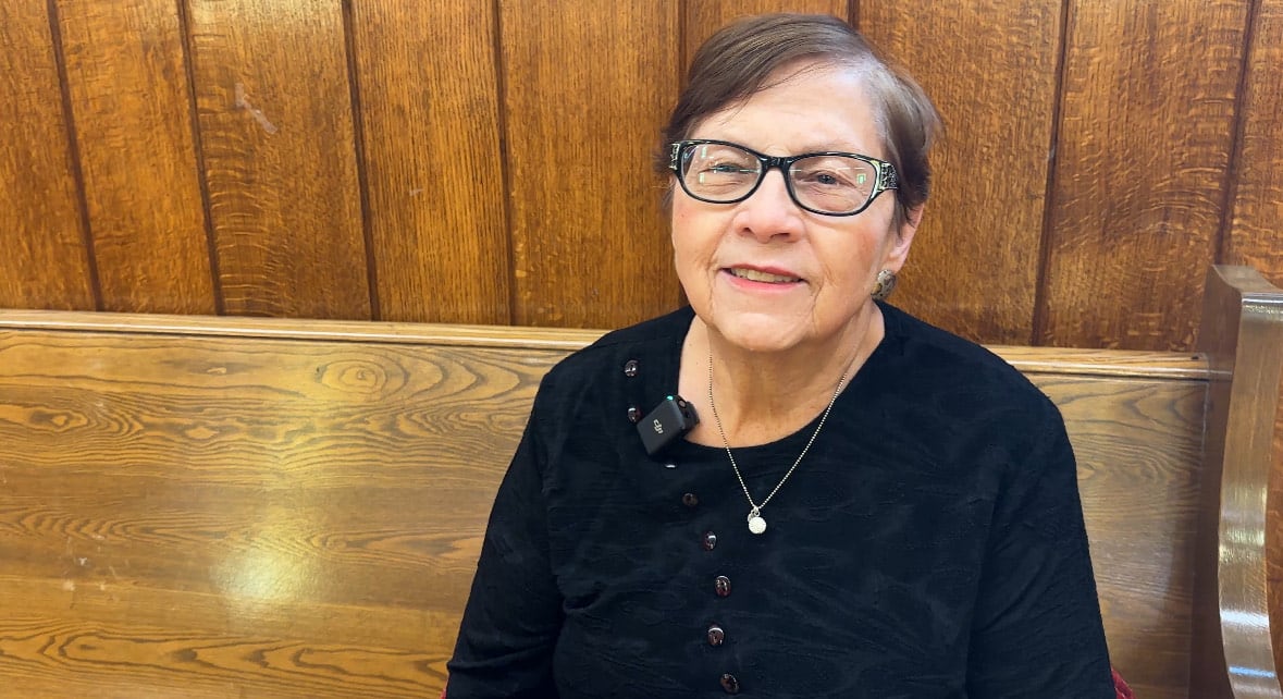A woman in a black shirt and glasses sits on a wooden bench.