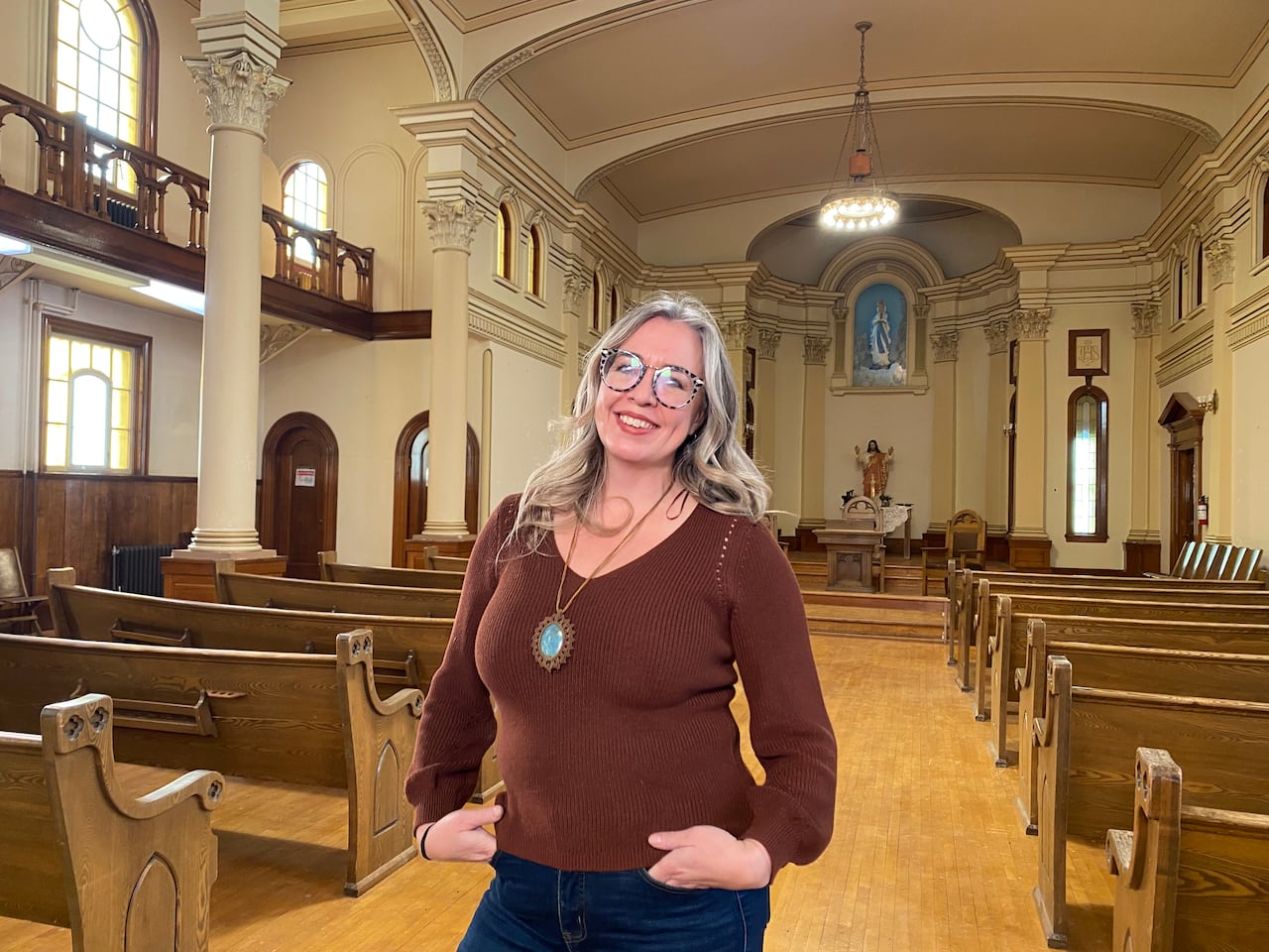 A woman in a brown V-neck sweater and jeans stands with her hands in her pockets, with a chapel space seen behind her.