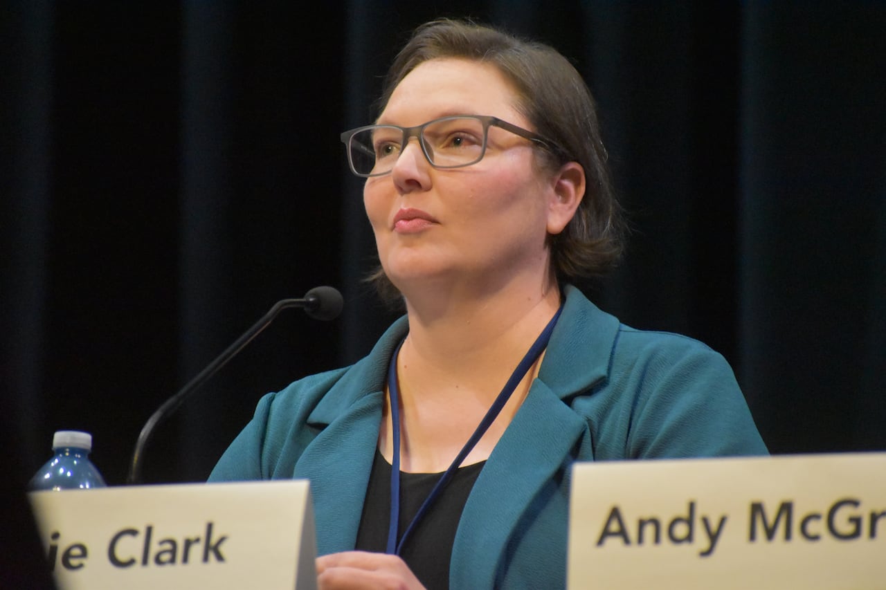 A woman with dark brown hair, light skin and glasses listens at a table with a dark cloth.
