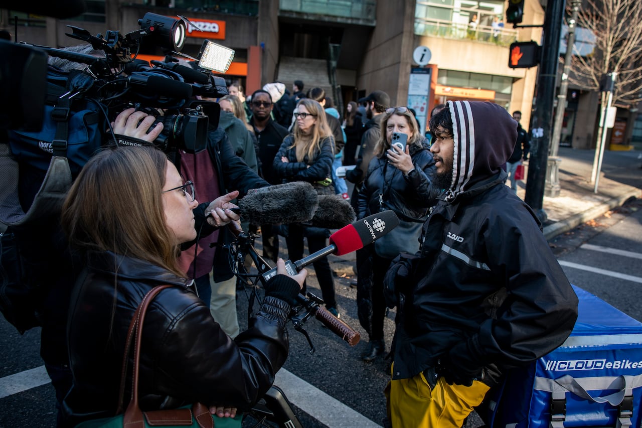 A man speaks into a microphone as people crowd around him and cameras film him.