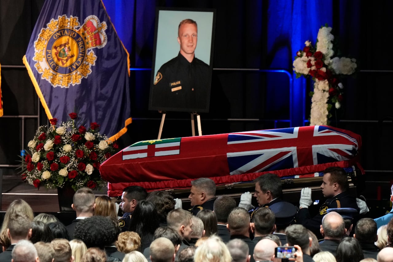 Several police officers carry a casket draped in a flag, in front of a stage where a photo of another police officer and a large bouquet of flowers sit.
