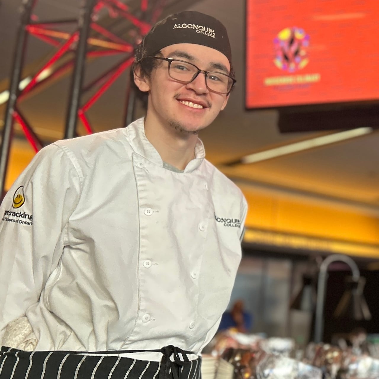 A young Inuk chef poses at an event wearing an apron that reads 'Algonquin College'