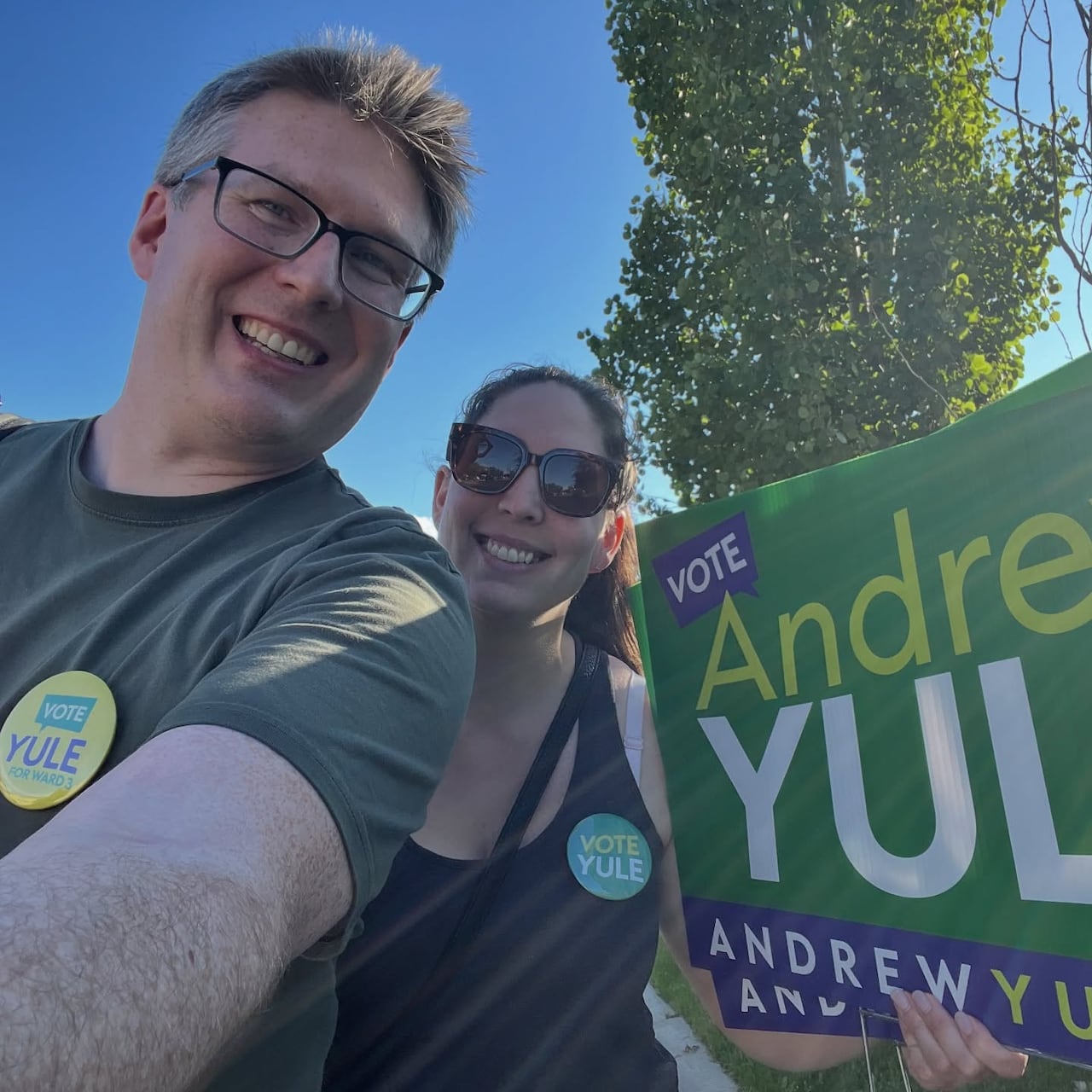A man with glasses smiles taking a selfie, with a woman holding an Andrew Yule campaign sign beside him
