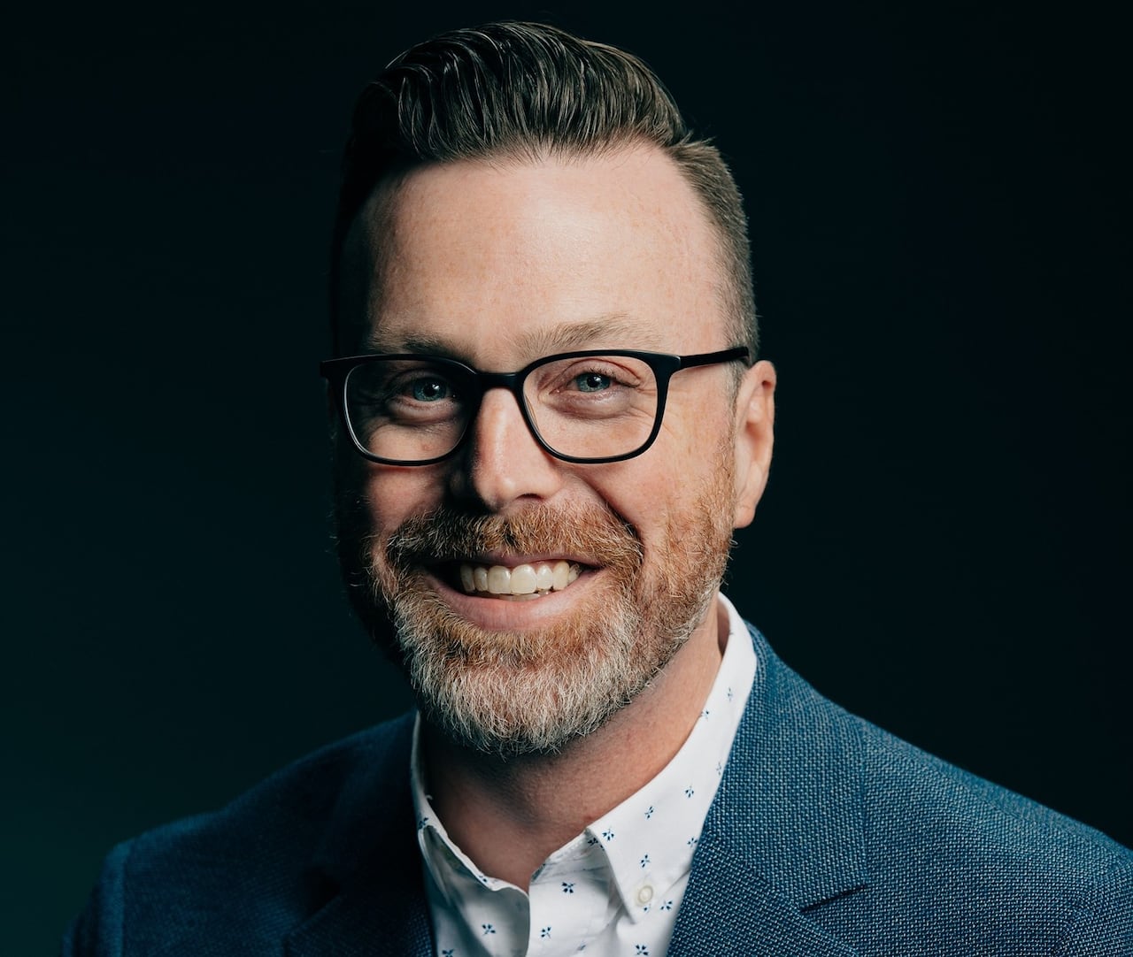A man with a blue suit and glasses smiles at the camera