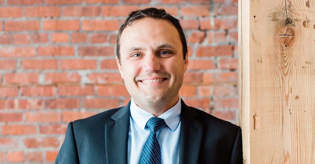 A man in a suit smiles at the camera and leans against a wooden post