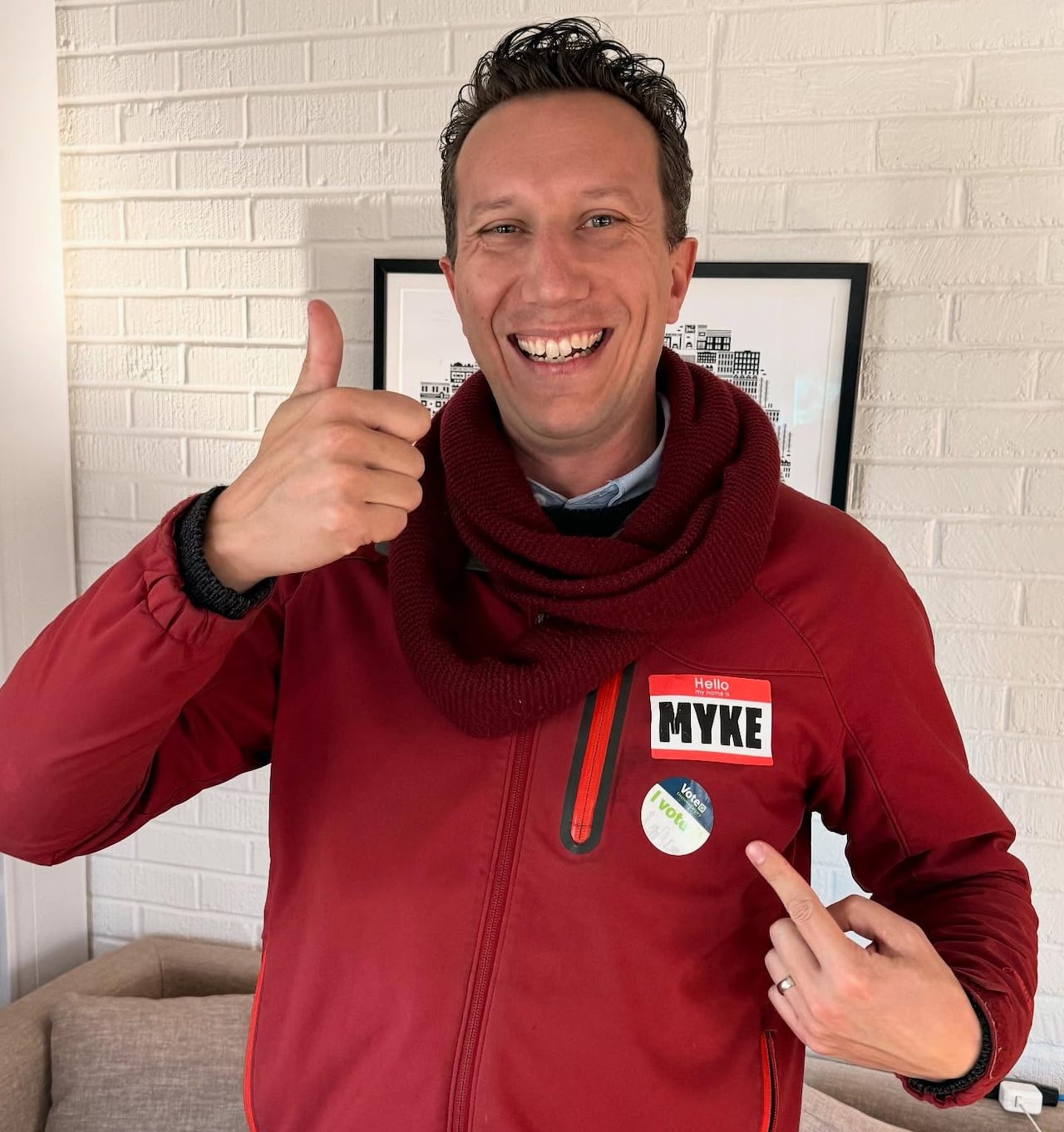 A man points at his nametag that says 'Myke' with a smile and a thumbs up 