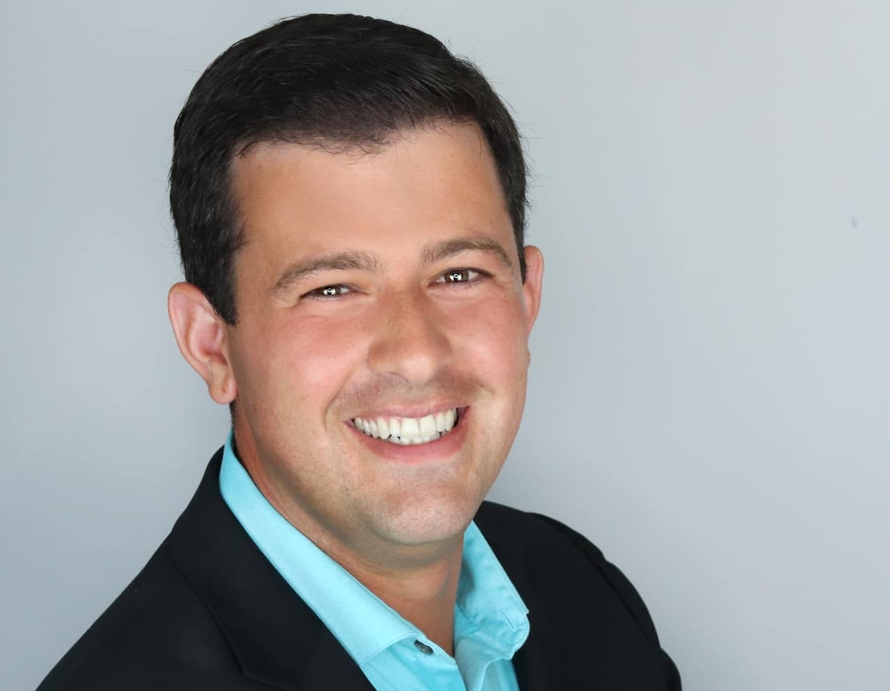 A man in a blue shirt and black suit jacket smiles at the camera