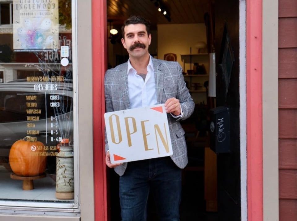 A man with a mustache leans in an open store doorway, holding a sign that says 'open'