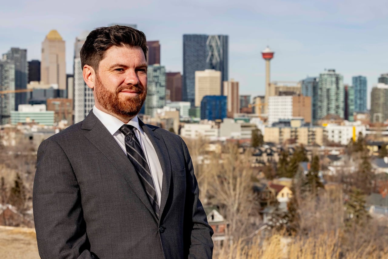 A man in a suit stands in front of the Calgary city skyline