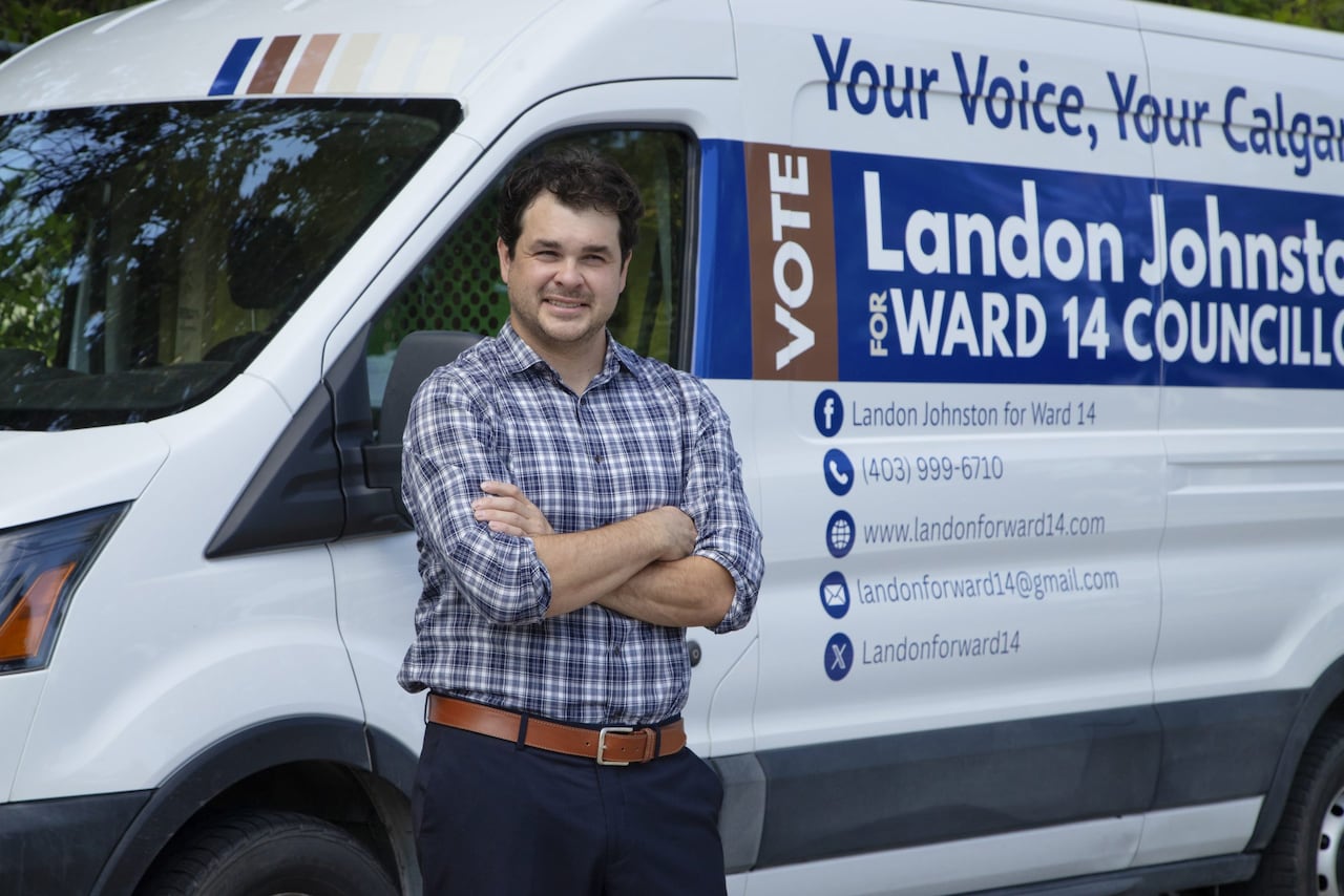 A man in a plaid shirt crosses his arms and leans against a white van with campaign information for Landon Johnston. 