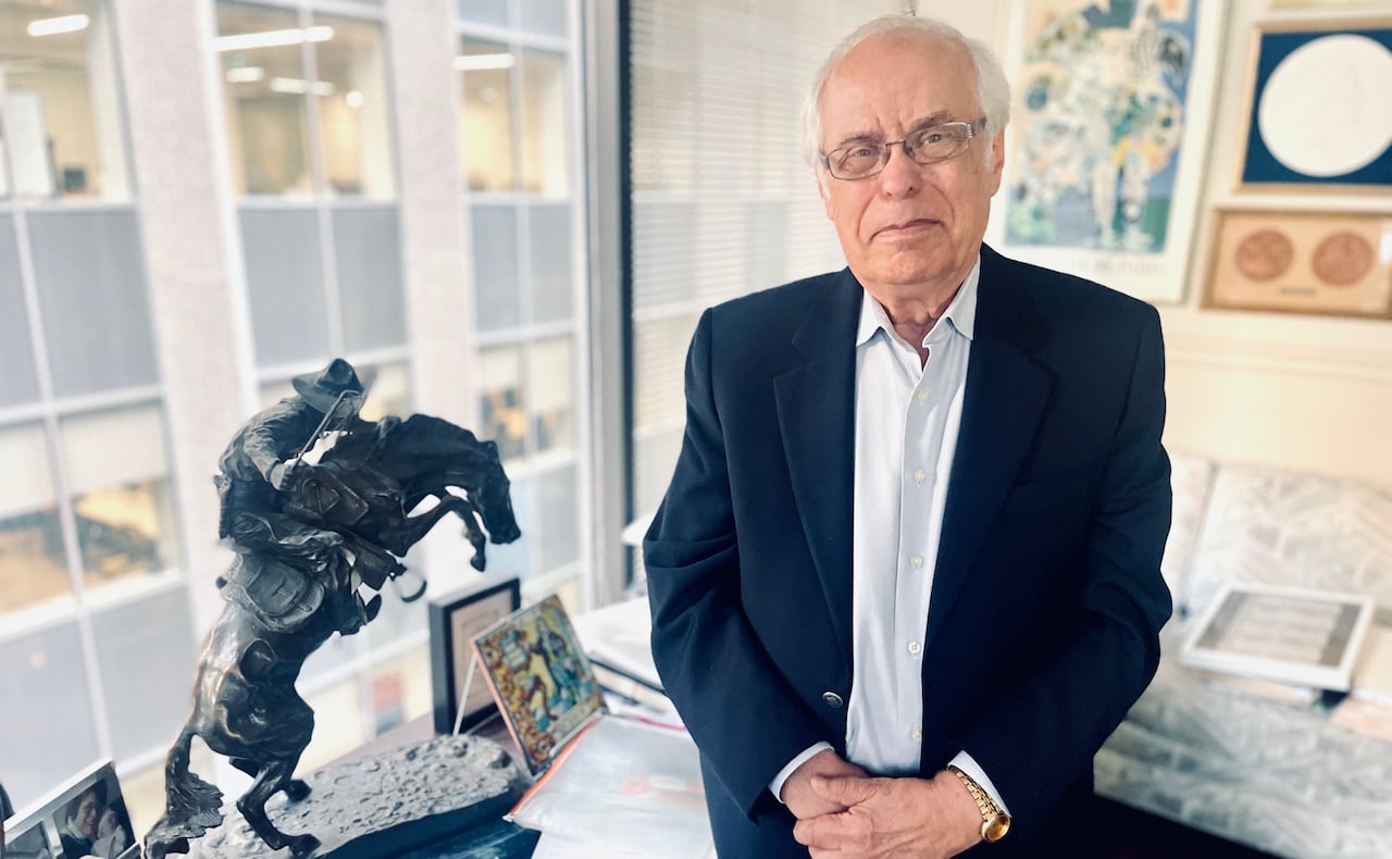 Bob Aaron, wearing a suit, stands nexts to his desk in front of his office window. 