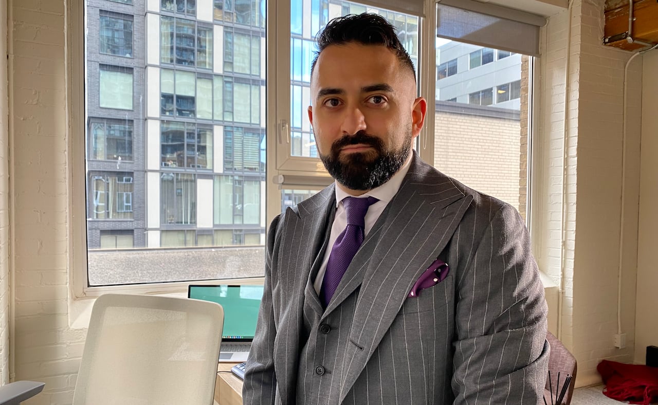 Justin Nasseri wearing a grey suit and purple tie stands in front of his desk with a window behind him. 