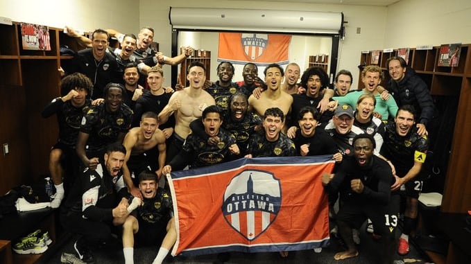 A group of men celebrate in a locker room and hold up a red, white and blue flag.