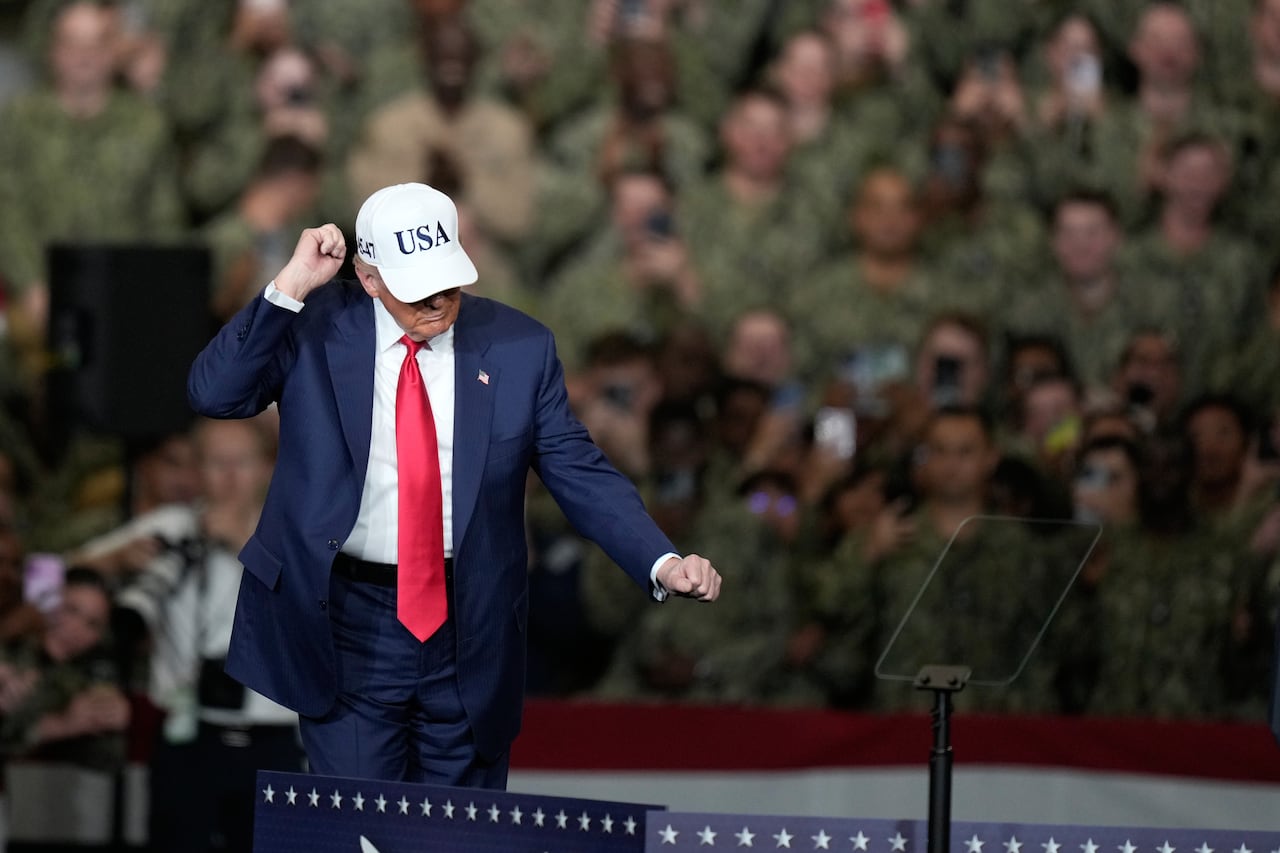 A man in a navy blue suit, with a red tie and a white ball cap with 'USA' on it, pumps his fist in the air while on a stage. Behind him is a crowd wearing military fatigues.