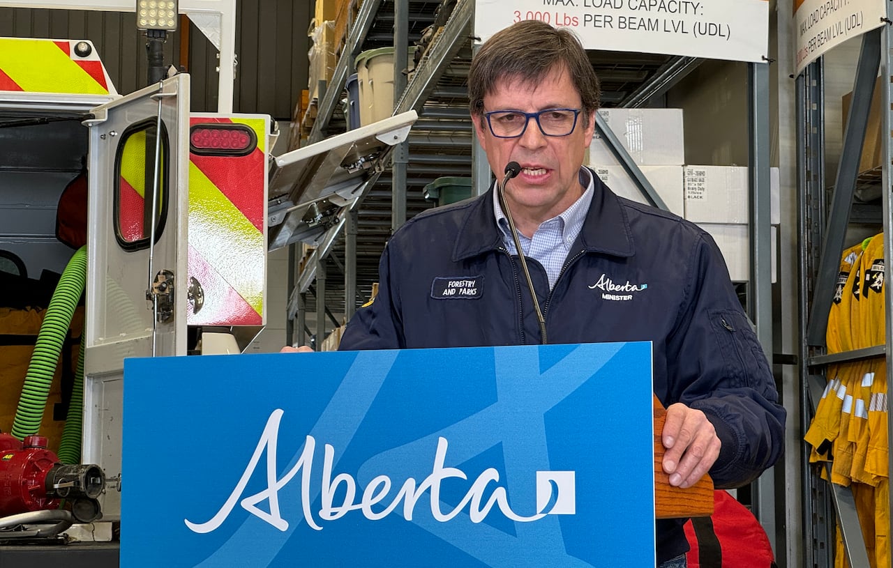 A man with glasses stands in front of a fire truck at a podium with a blue sign that says "Alberta."