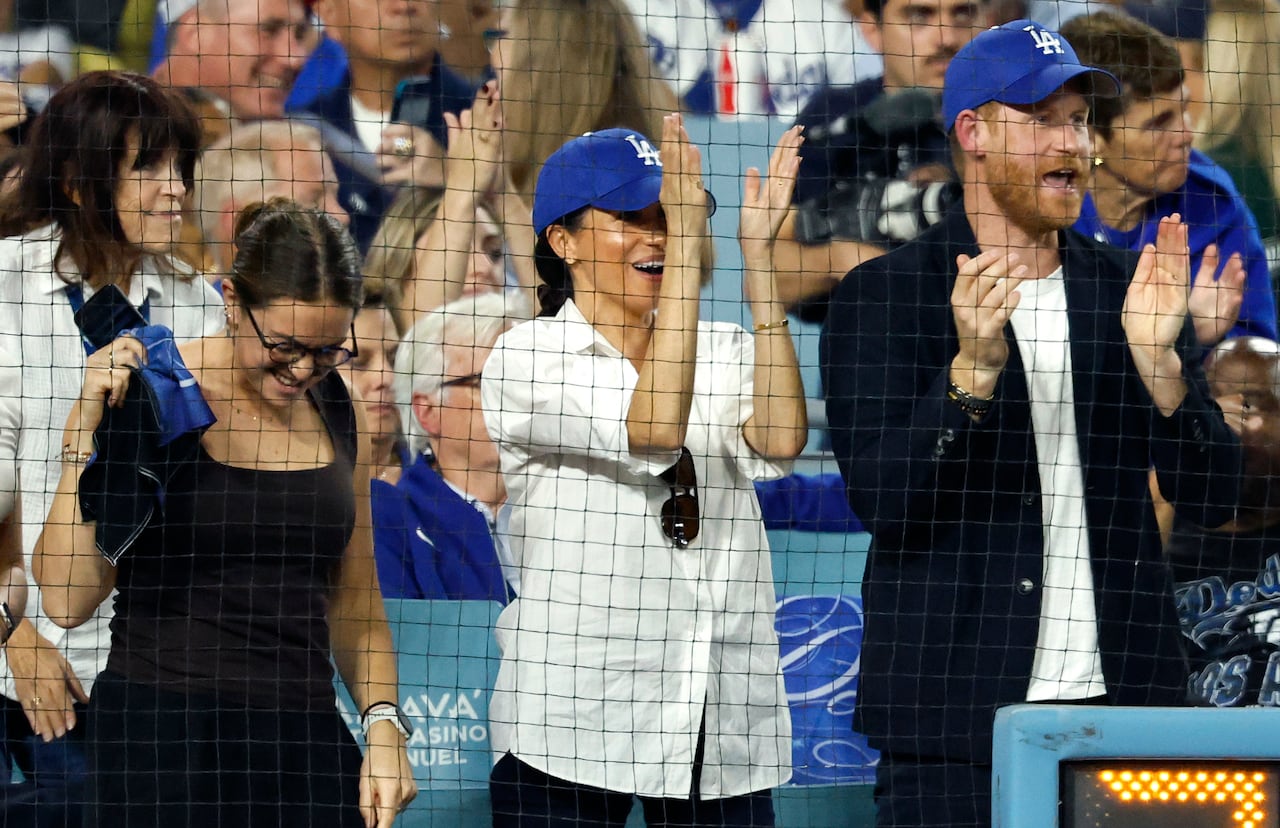 Meghan Markle in a white baseball jersey and blue ball cap stands next to Prince Harry in navy sports jacket, white t-shirt and blue ballcap as the clap and cheer in the stands of a baseball stadium