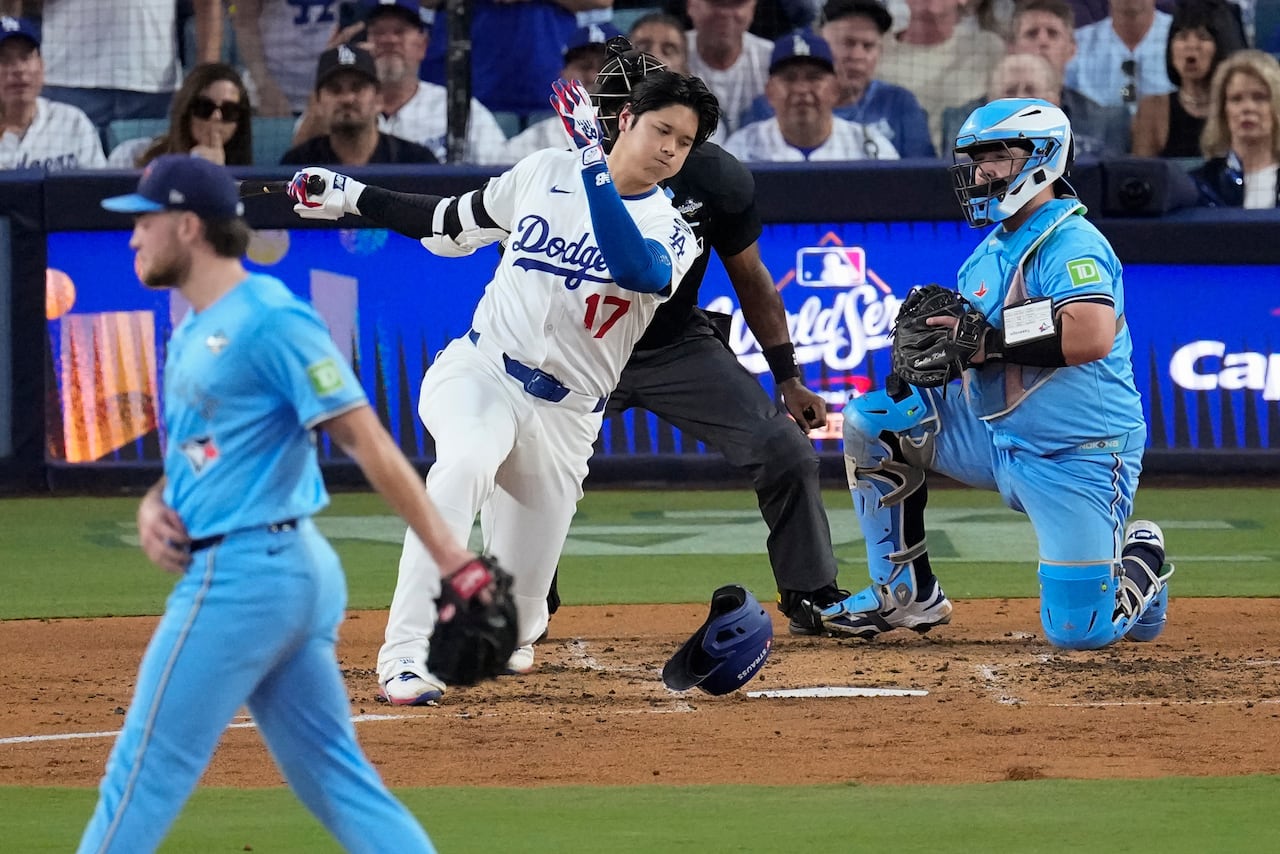 A baseball player swings, misses and loses his helmet as a catcher and umpire look on. The pitcher waits on the mound. 