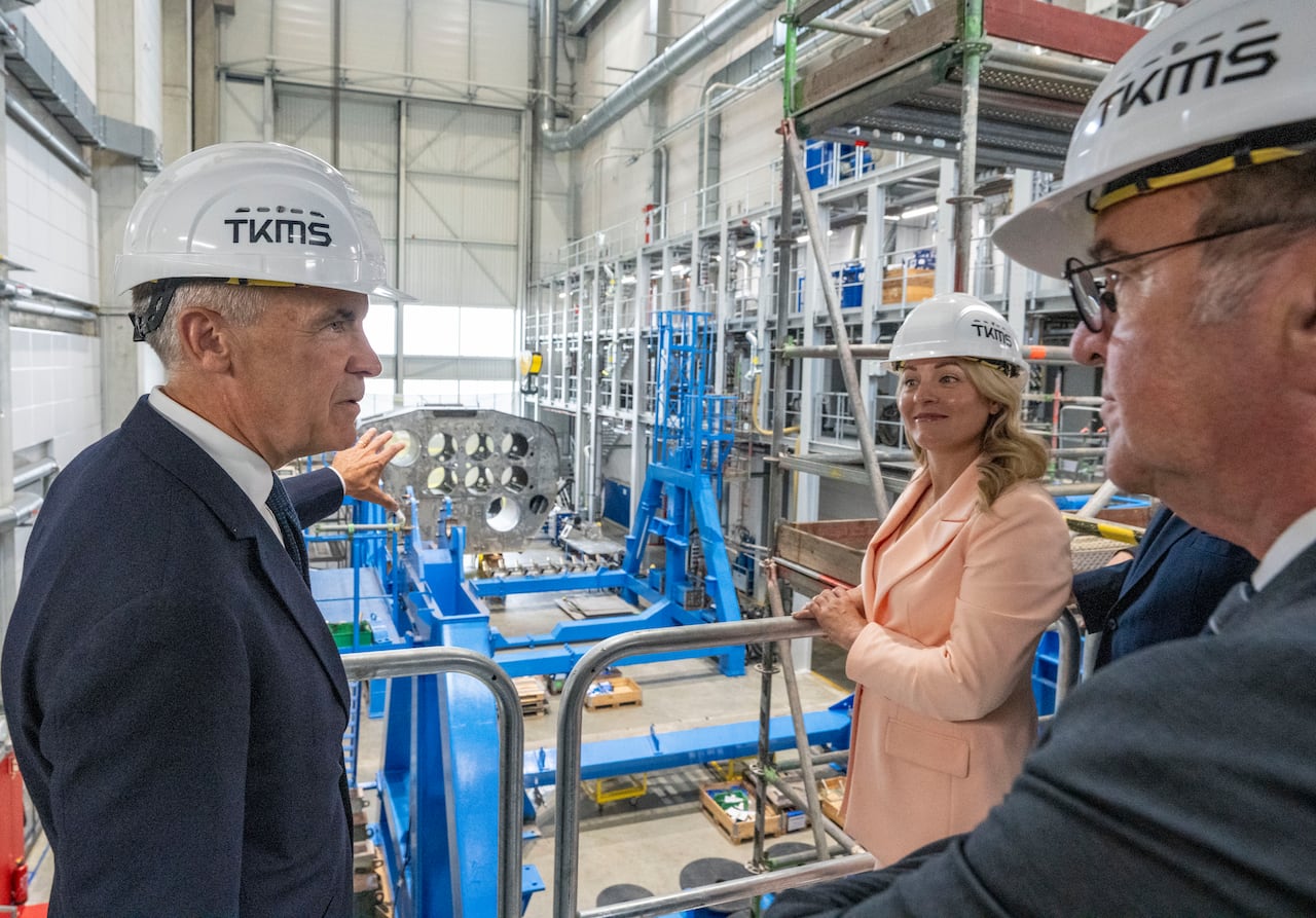 Two men and a woman wearing hard hats point to equipment in a factory.