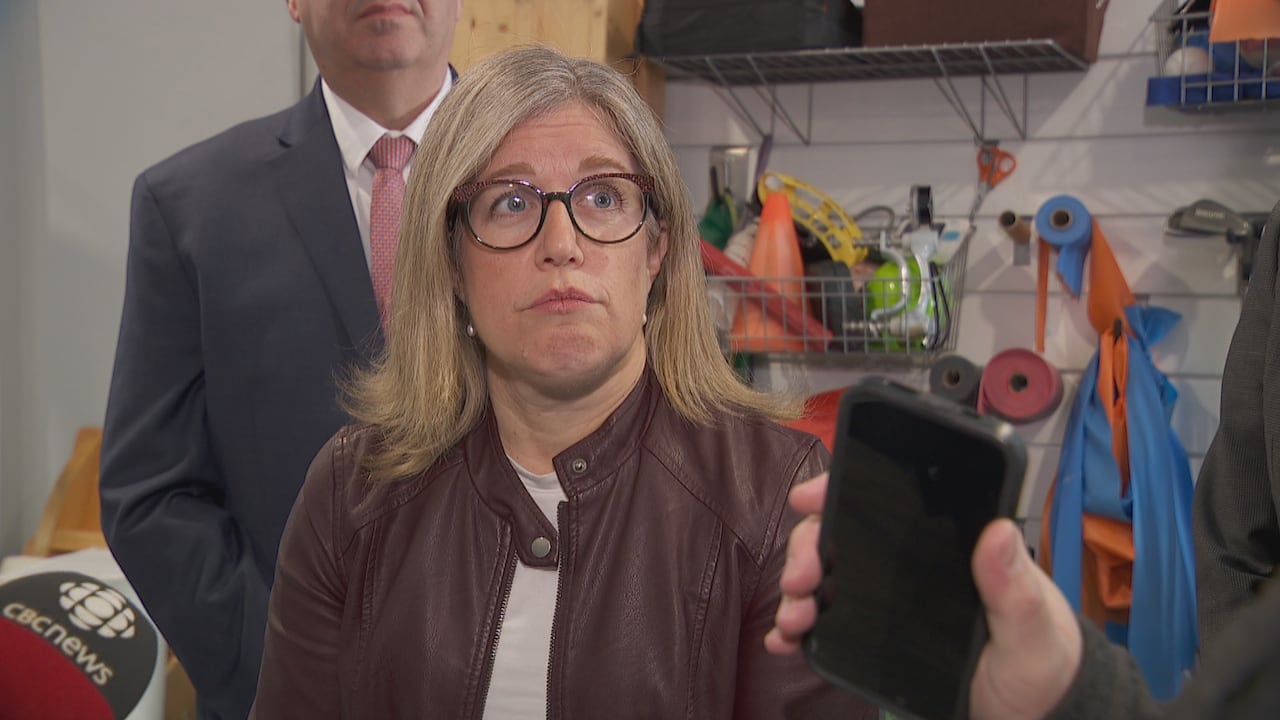 A blond woman with glasses wearing a leather jacket looks to the right. Microphones and a cell phone are in the foreground. 