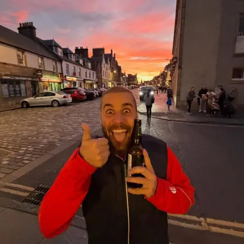 Justin Bieber Justin Bieber gives a thumbs up sign while holding a beer outside. A cobbled street and shops are seen in the background
