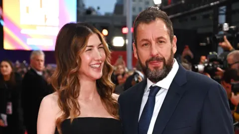 Getty Images Jackie Sandler smiling at her husband Adam Sandler, as he poses on the red carpet