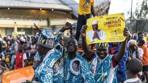 MARCO LONGARI/AFP via Getty Images Three men, smiling and gesturing, dressed in bright and pattered matching blue outfits, hold up a sign in support of the opposition leader. There are many people behind them, in a rally.
