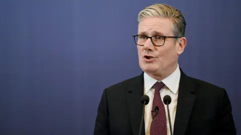 EPA Sir Keir Starmer, dressed in a dark suit, white shirt, and red patterned tie, stands at a podium speaking during a news conference in Mumbai. Two microphones are positioned in front of him, and the backdrop is a plain blue screen.