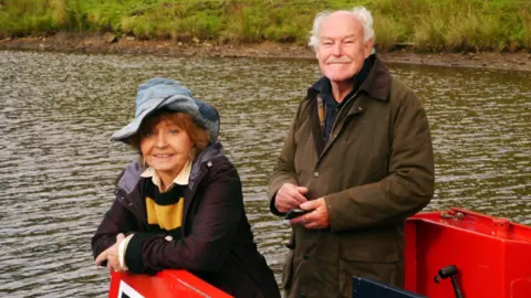Alamy Prunella Scales and Timothy West in Great Canal Journeys. The couple are shown looking at camera from a lock on a canal.  Scales is wearing a floppy blue hat.