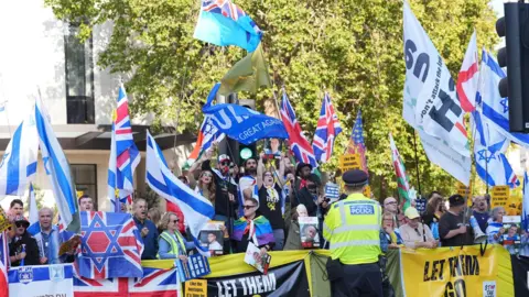 PA Media A crowd waving various flags stands behind a low fence row of banners, as a police officer with his back to the camera watches on. Many hold Israeli flags aloft, while some union jacks and St George's flags can also be seen. The crowd consists of men and women, and some are shouting. 